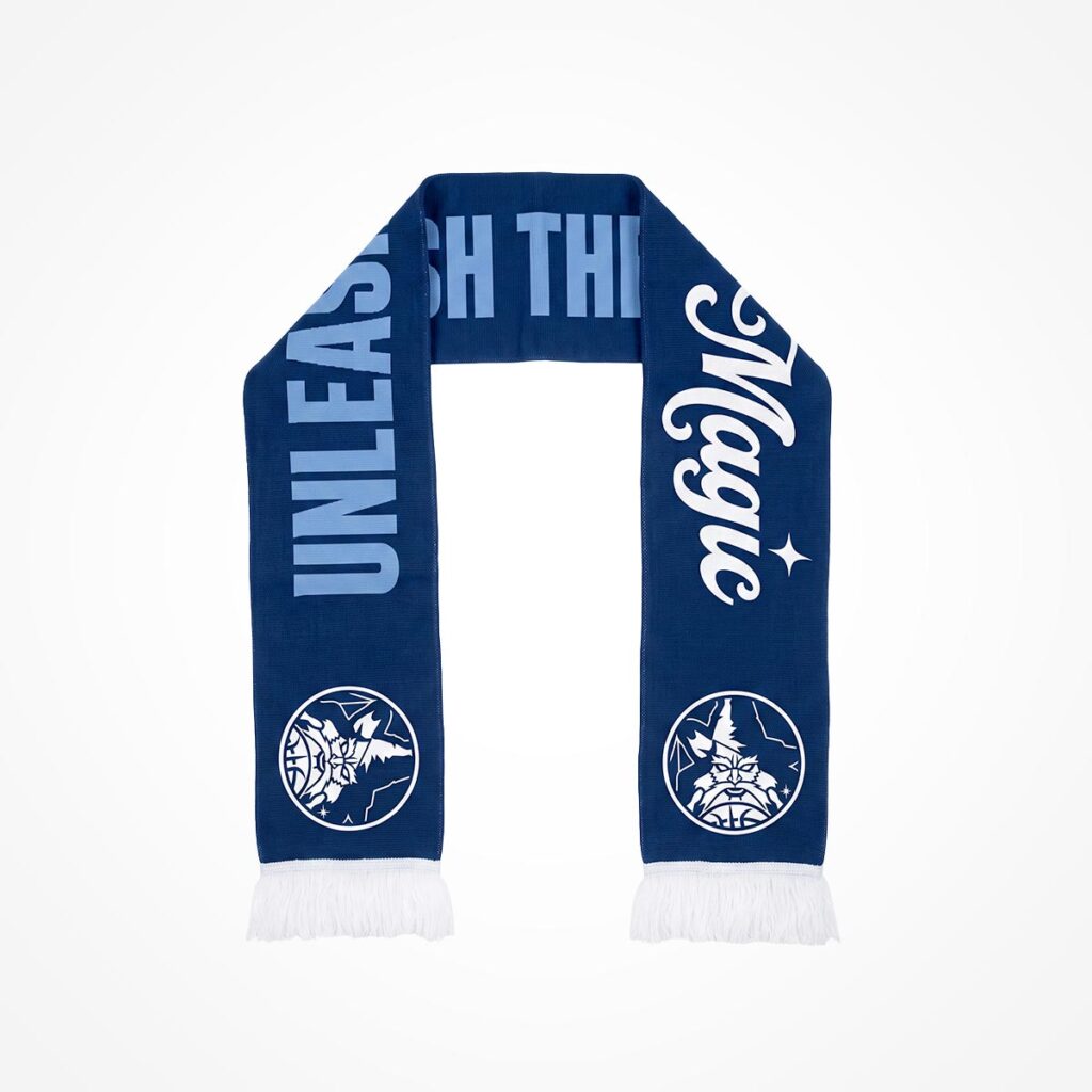 Ein blauer Schal mit der weißen Aufschrift „UNLEASH THE MAGIC“. An beiden Enden befindet sich ein weißes Logo mit dem Kopf der Freiheitsstatue, und die Enden sind mit weißen Fransen verziert.