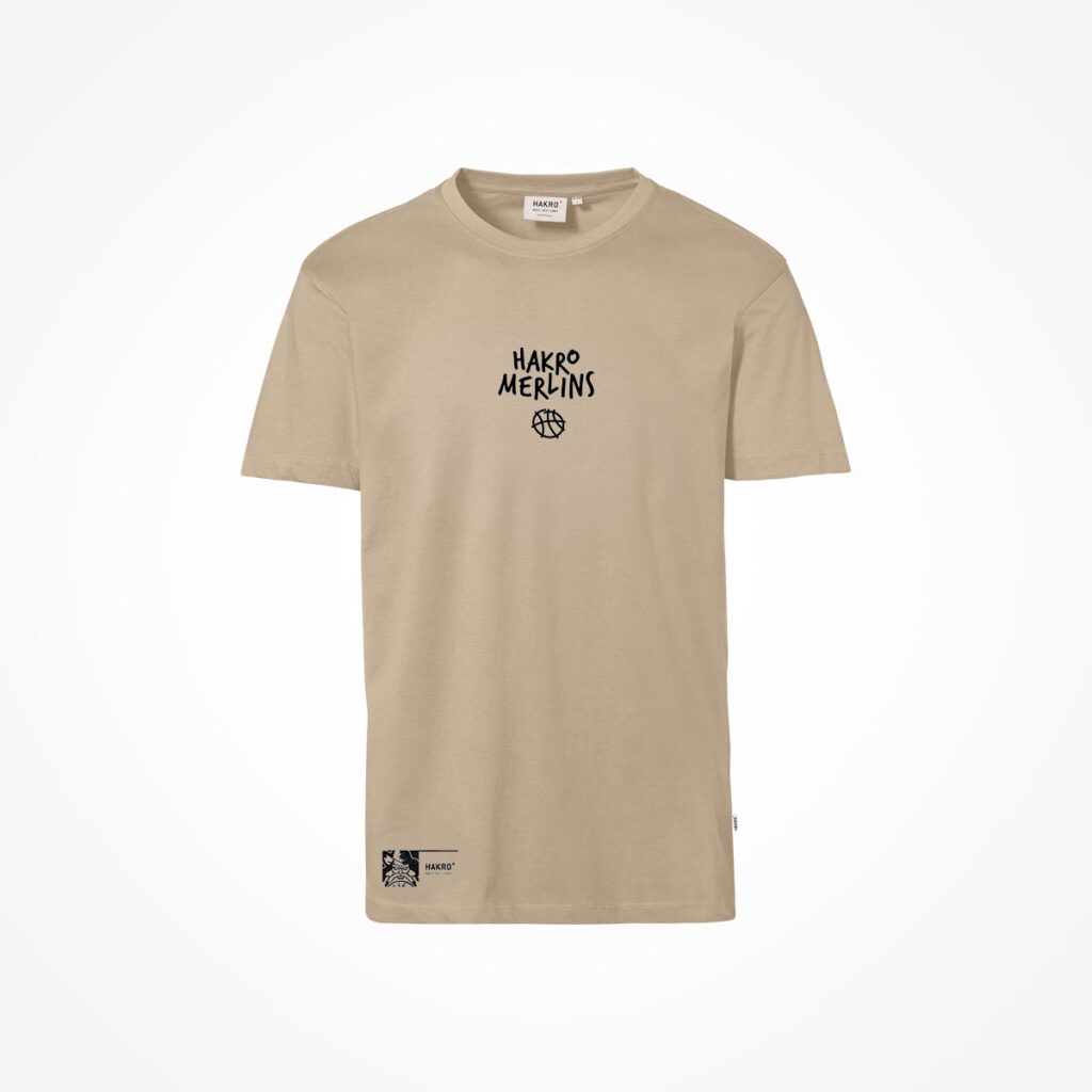 Beiges T-Shirt mit kurzen Ärmeln, mit dem Aufdruck „HAKRO MERLINS“ und einer kleinen Basketball-Grafik auf der Brust. Das Shirt hat ein kleines schwarz-weißes Etikett nahe dem unteren Saum.