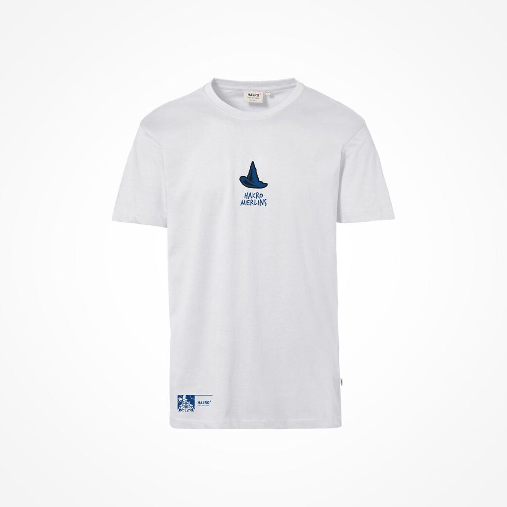 Weißes T-Shirt mit einem blauen Aufdruck, der einen Hexenhut und den Schriftzug „MADE MECINIS“ auf der Brust zeigt. Außerdem befindet sich unten links am Saum ein kleiner blau-weißer rechteckiger Aufnäher.